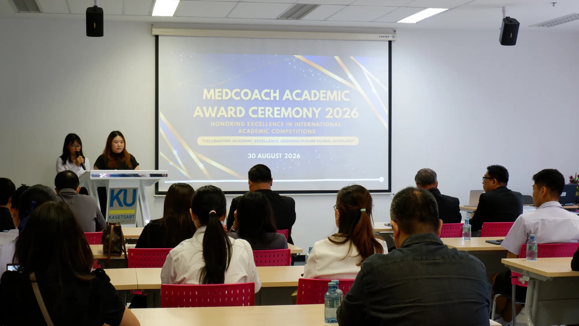 Two hosts open the MedCoach Academic Award Ceremony 2026 at a lectern in front of a seated audience, with the opening slide on the screen behind them.