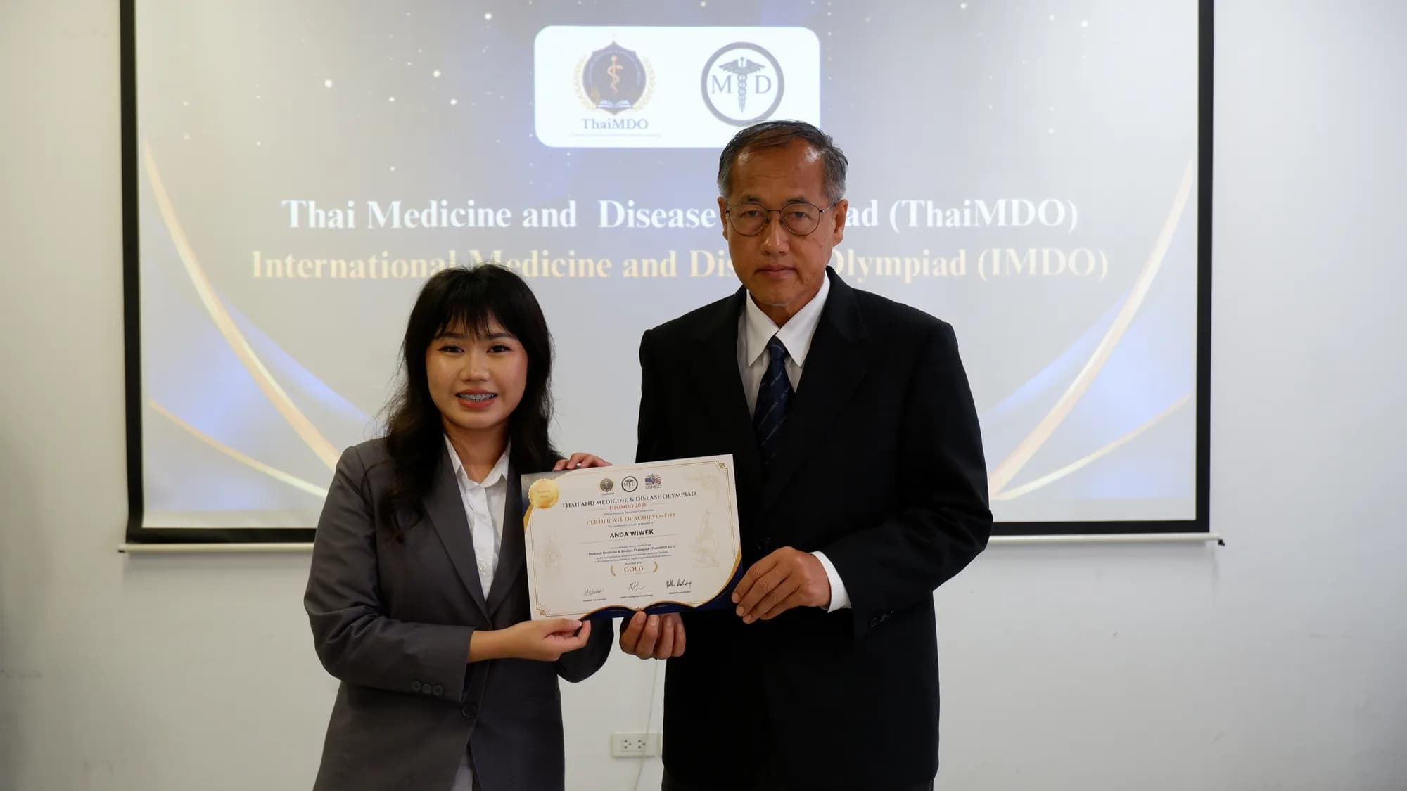 A student receives a certificate from a committee member, with the ThaiMDO and IMDO slide on the screen behind.