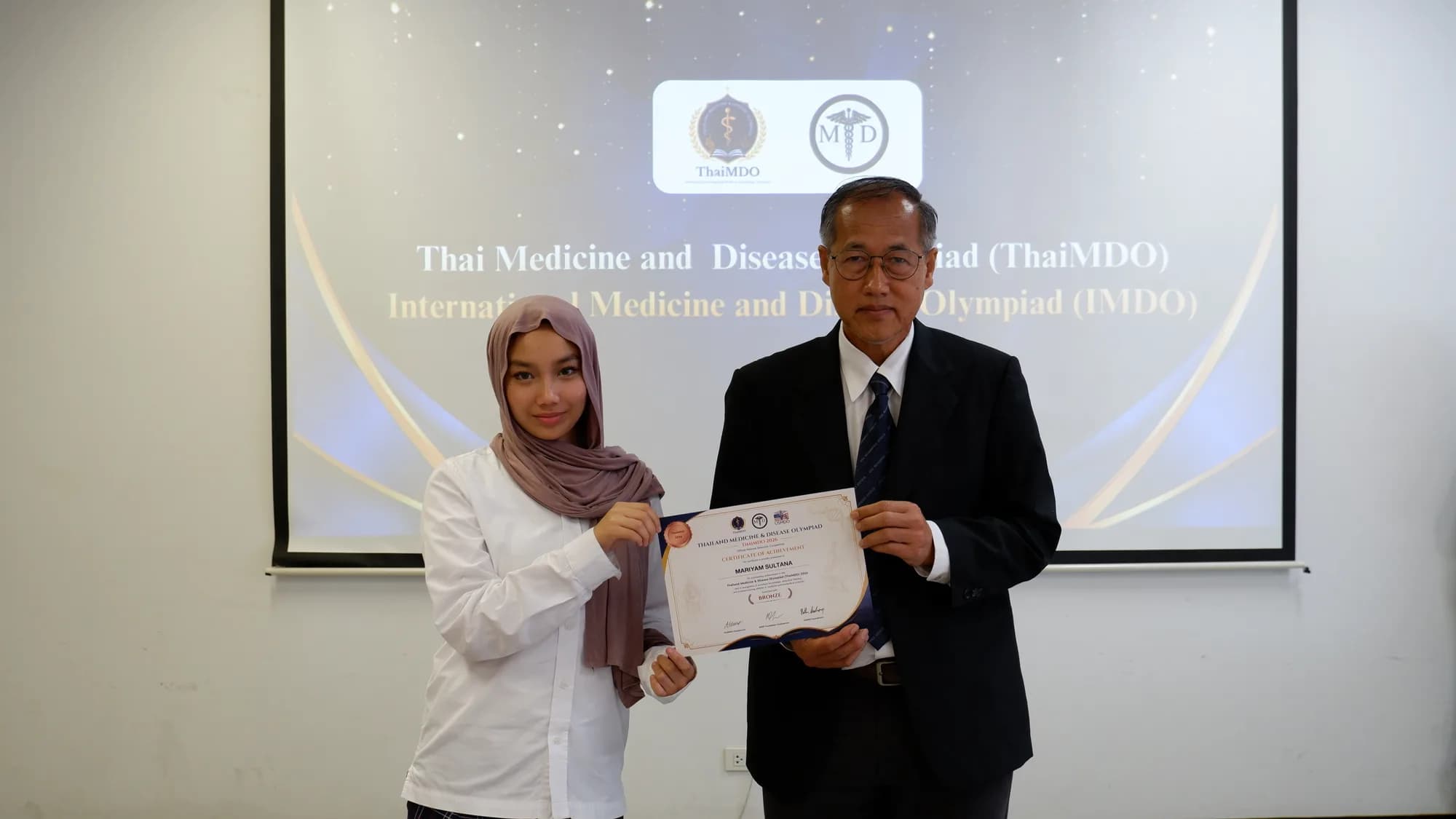 A student wearing a hijab receives a certificate from a committee member, with the ThaiMDO and IMDO slide on the screen behind.