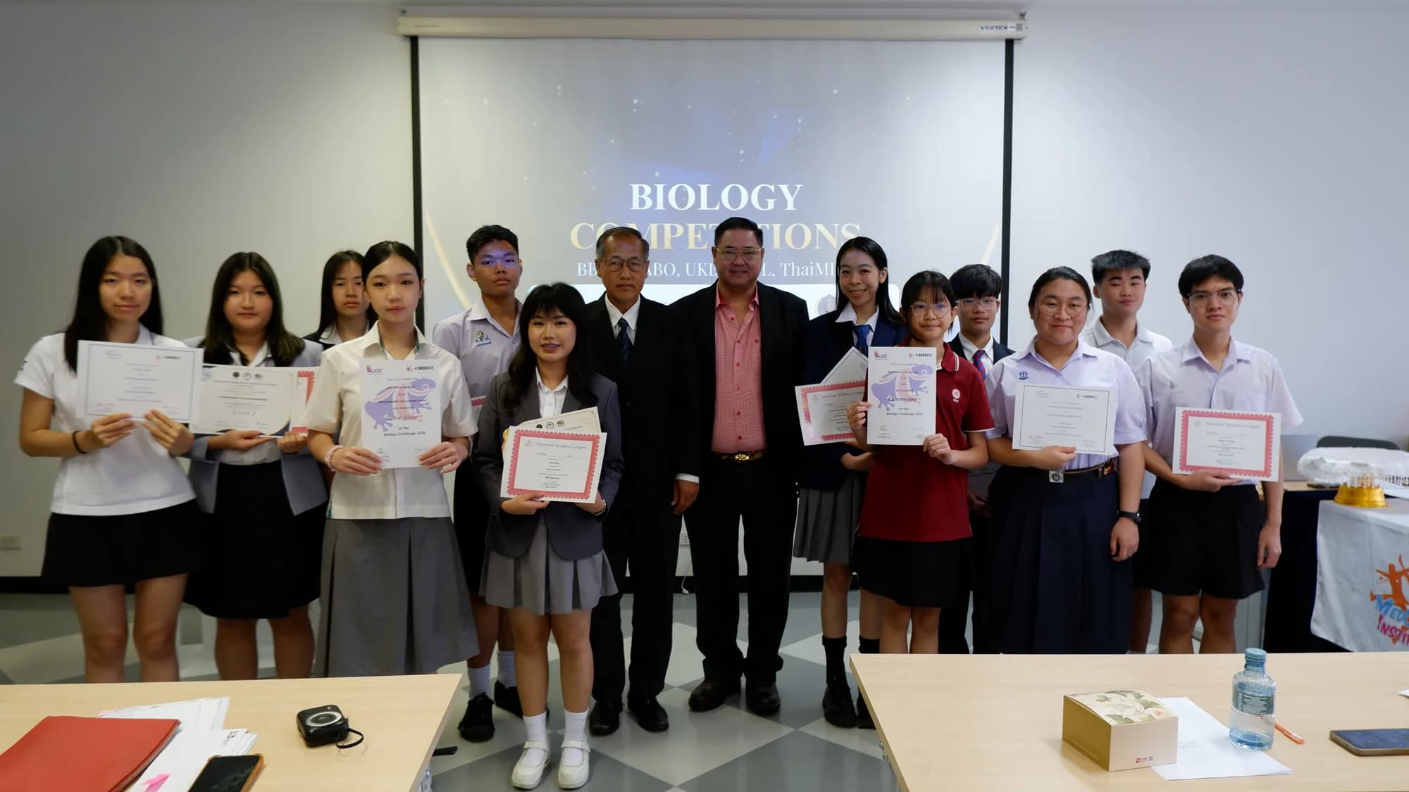 About twelve students hold their certificates in a group photograph alongside two committee members, with a biology competitions slide on the screen behind.