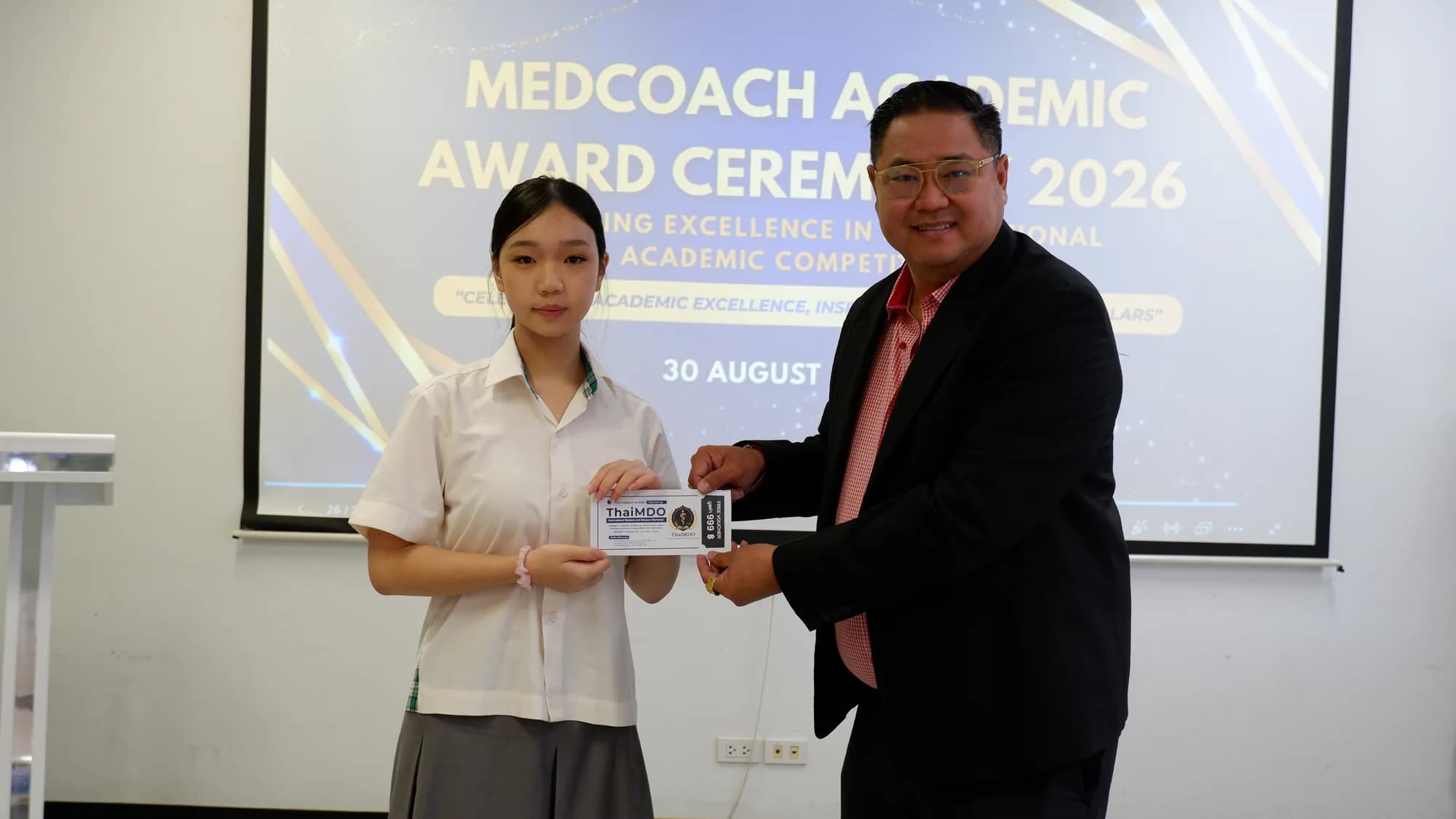 A student receives a ThaiMDO award plaque from a committee member, with the ceremony title slide on the screen behind.
