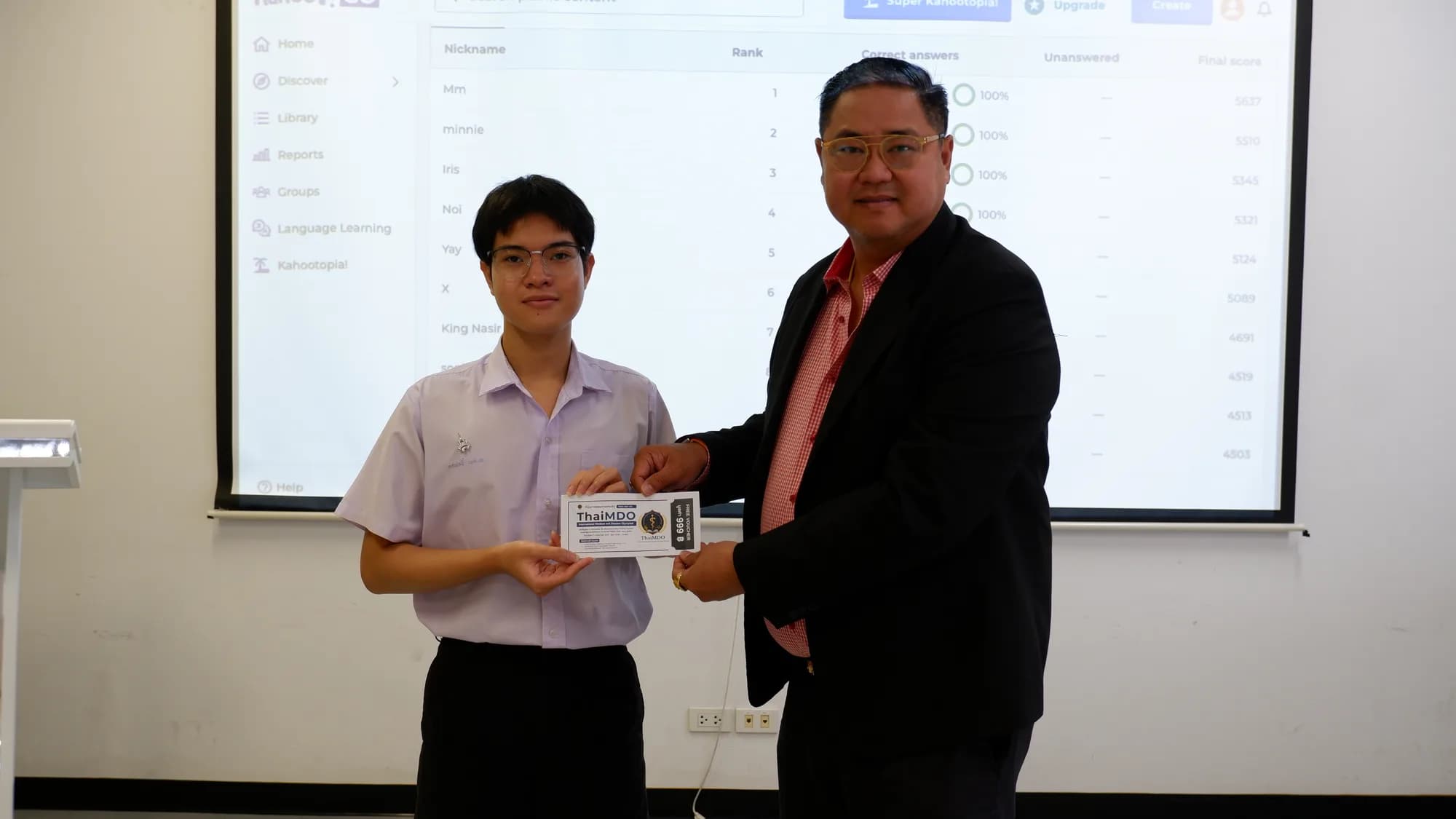 A student receives a ThaiMDO award plaque with the results leaderboard displayed on the screen behind.